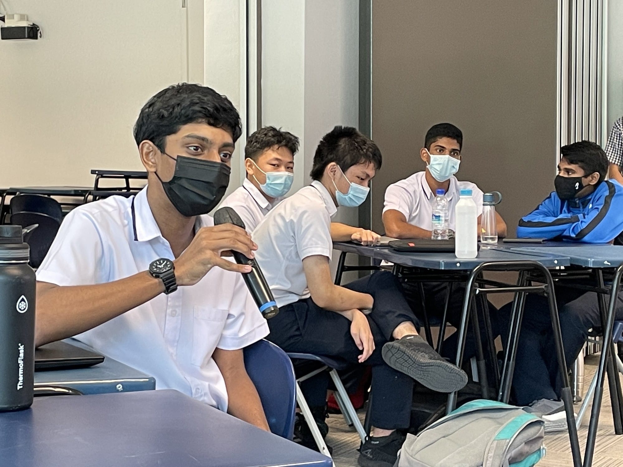 Five masked male students in school uniforms sit at desks; one holds a microphone and speaks.